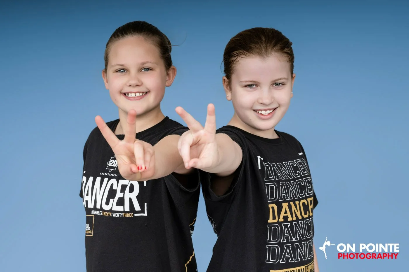 Two young girls in black dance shirts making peace signs against a blue background.