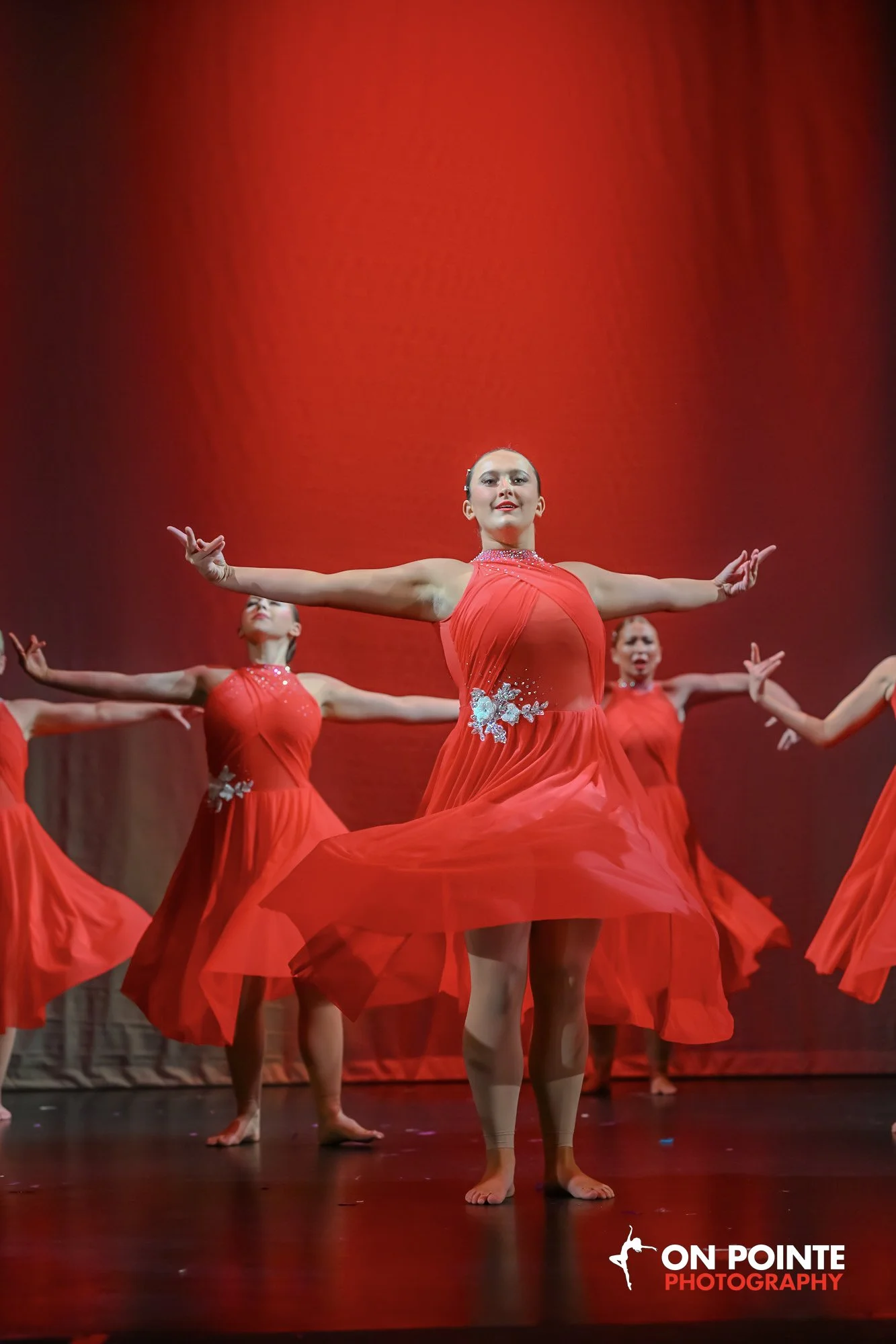 A group of female dancers performing on stage, wearing red dresses with floral embellishments, against a red background. The lead dancer is positioned in the center with her arms extended outward.