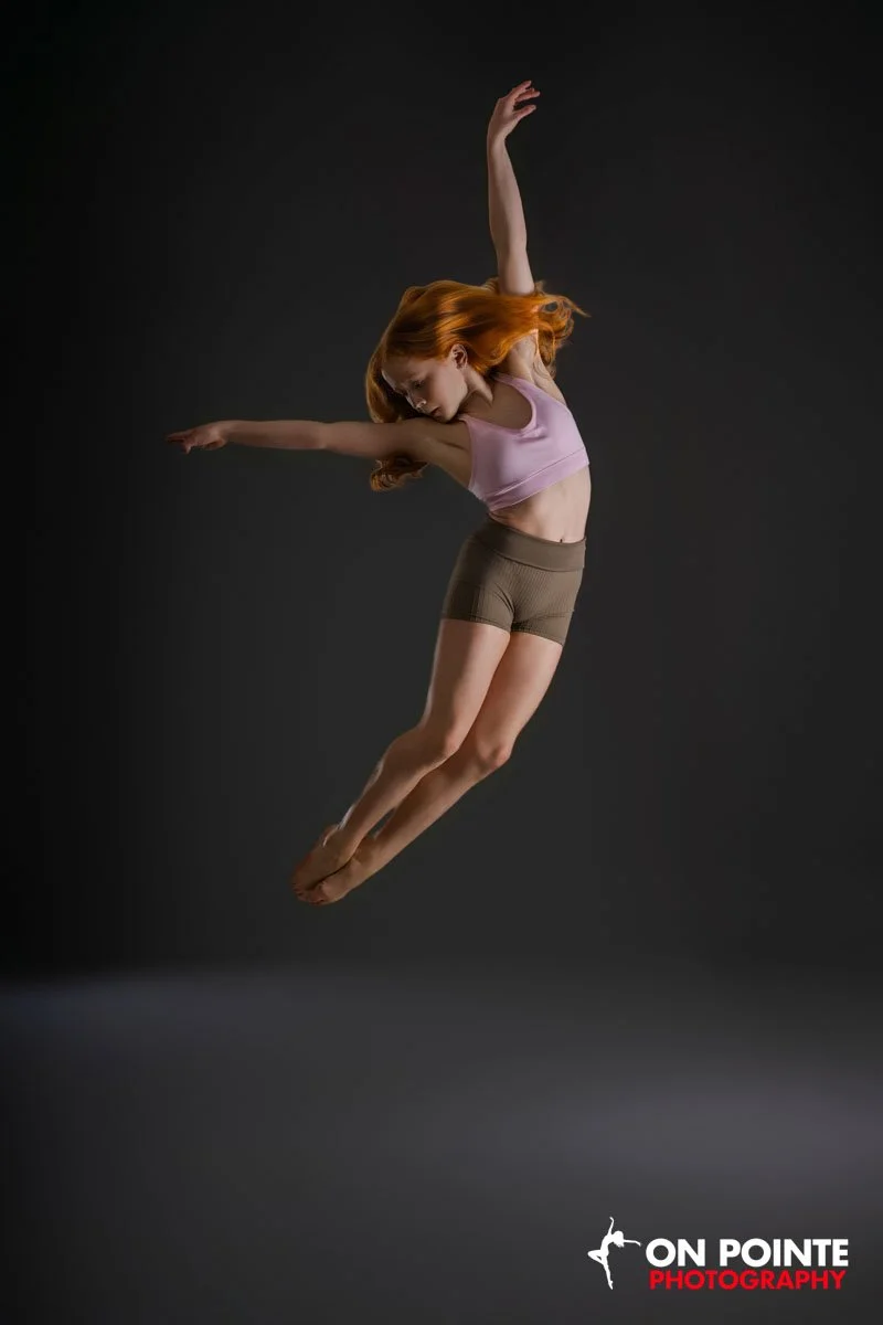 A woman with long red hair wearing a pink sports bra and brown shorts is captured mid-air in a dance or acrobatic pose against a dark background.