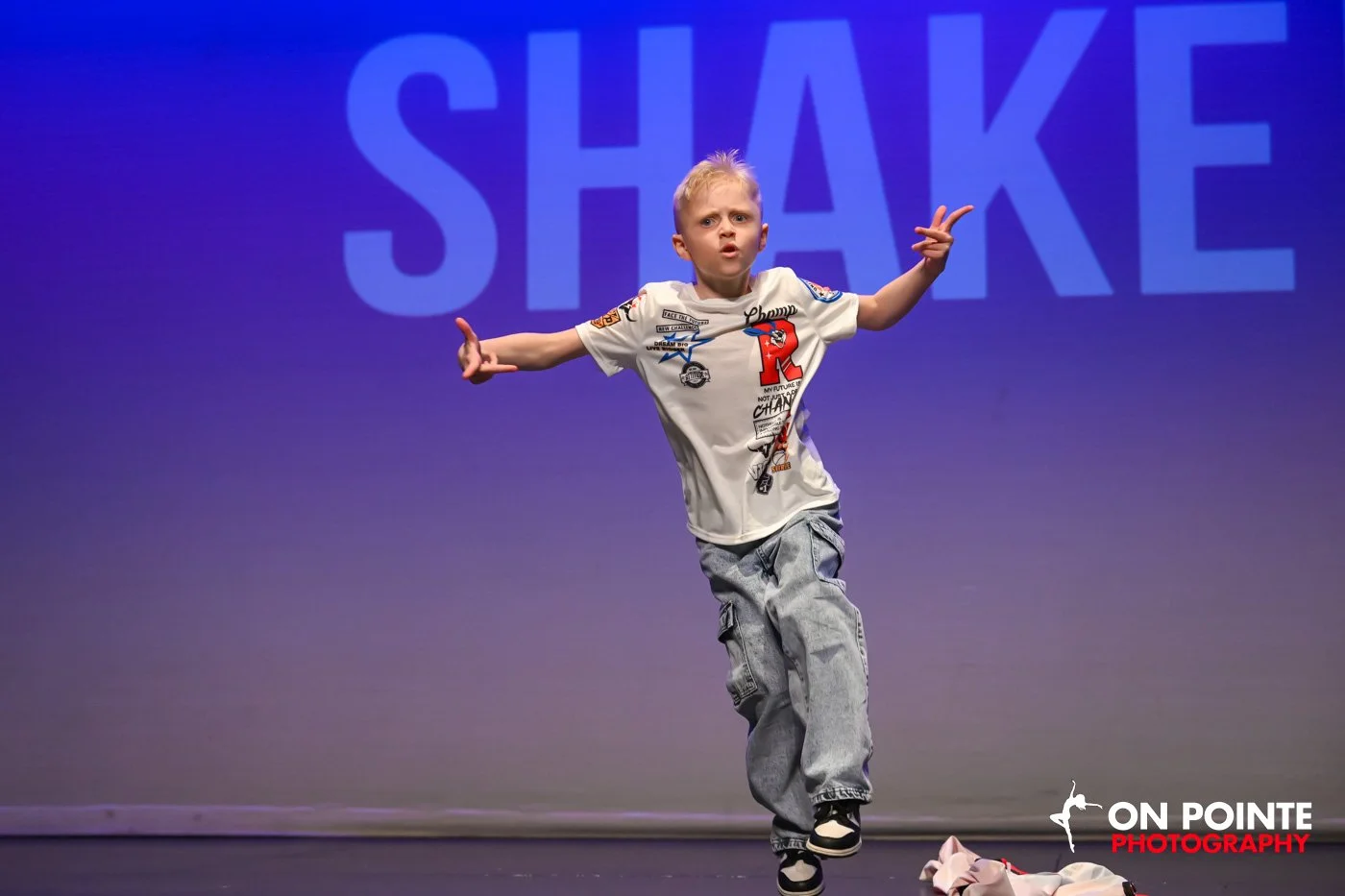 A young boy with blonde hair wearing a white graphic T-shirt, jeans, and sneakers performing a dance move on stage, with a large blue background displaying the word 'SHAKE' behind him and the logo 'ON POINTE PHOTOGRAPHY' in the bottom right corner.