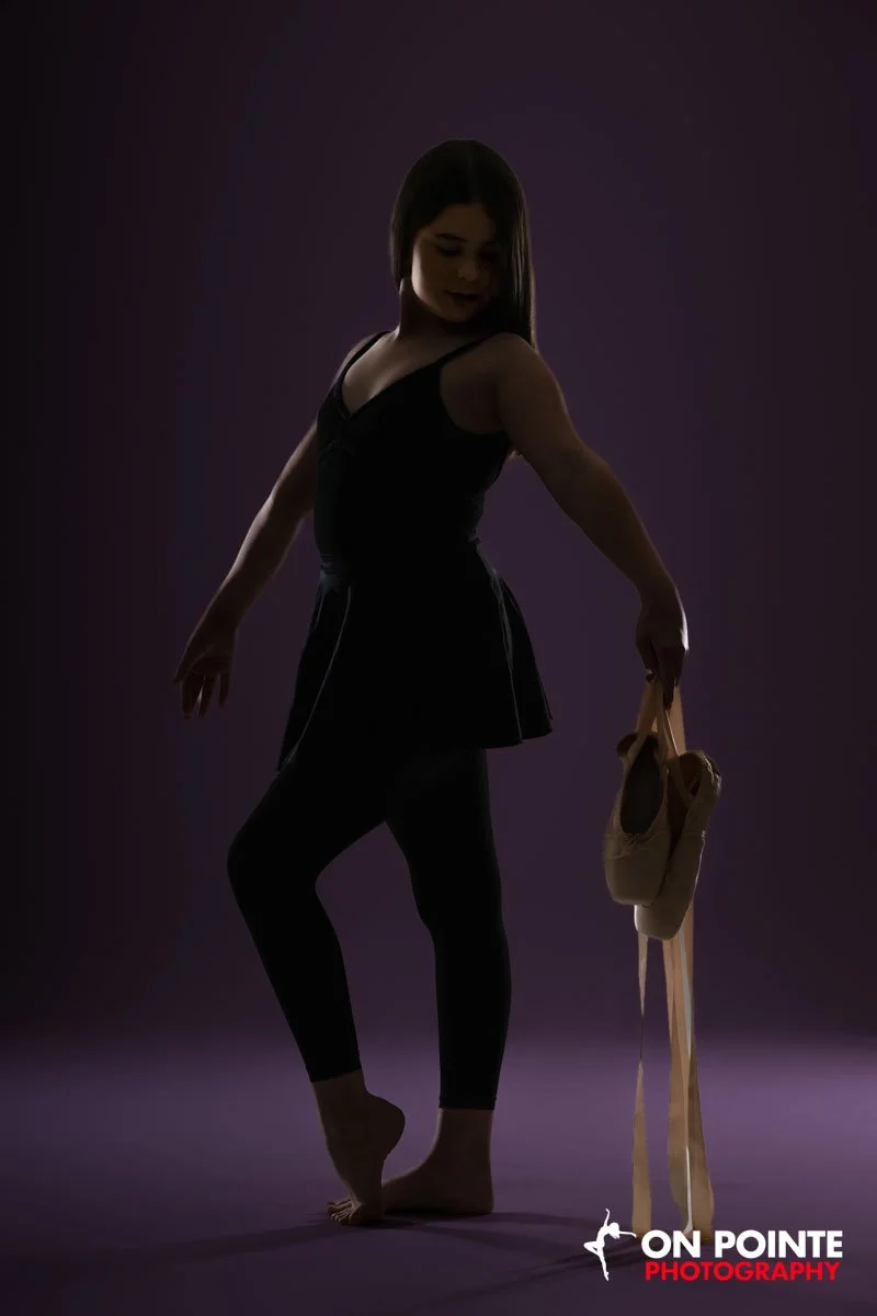 A ballet dancer stands on one foot in a dark studio, holding ballet shoes in her hand, with the ON POINTE PHOTOGRAPHY logo in the corner.
