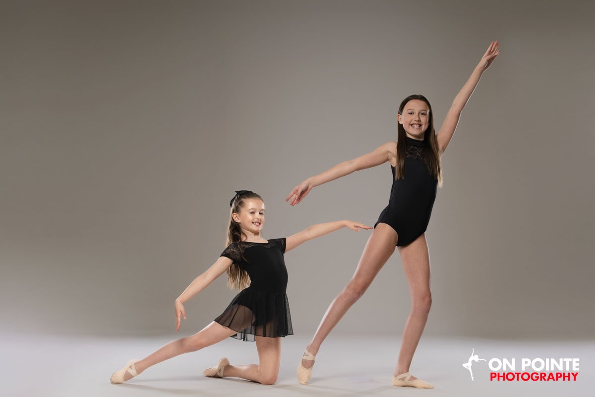 Two young girls in ballet poses wearing black leotards, one younger and one older, dancing on a studio floor with a plain grey background.