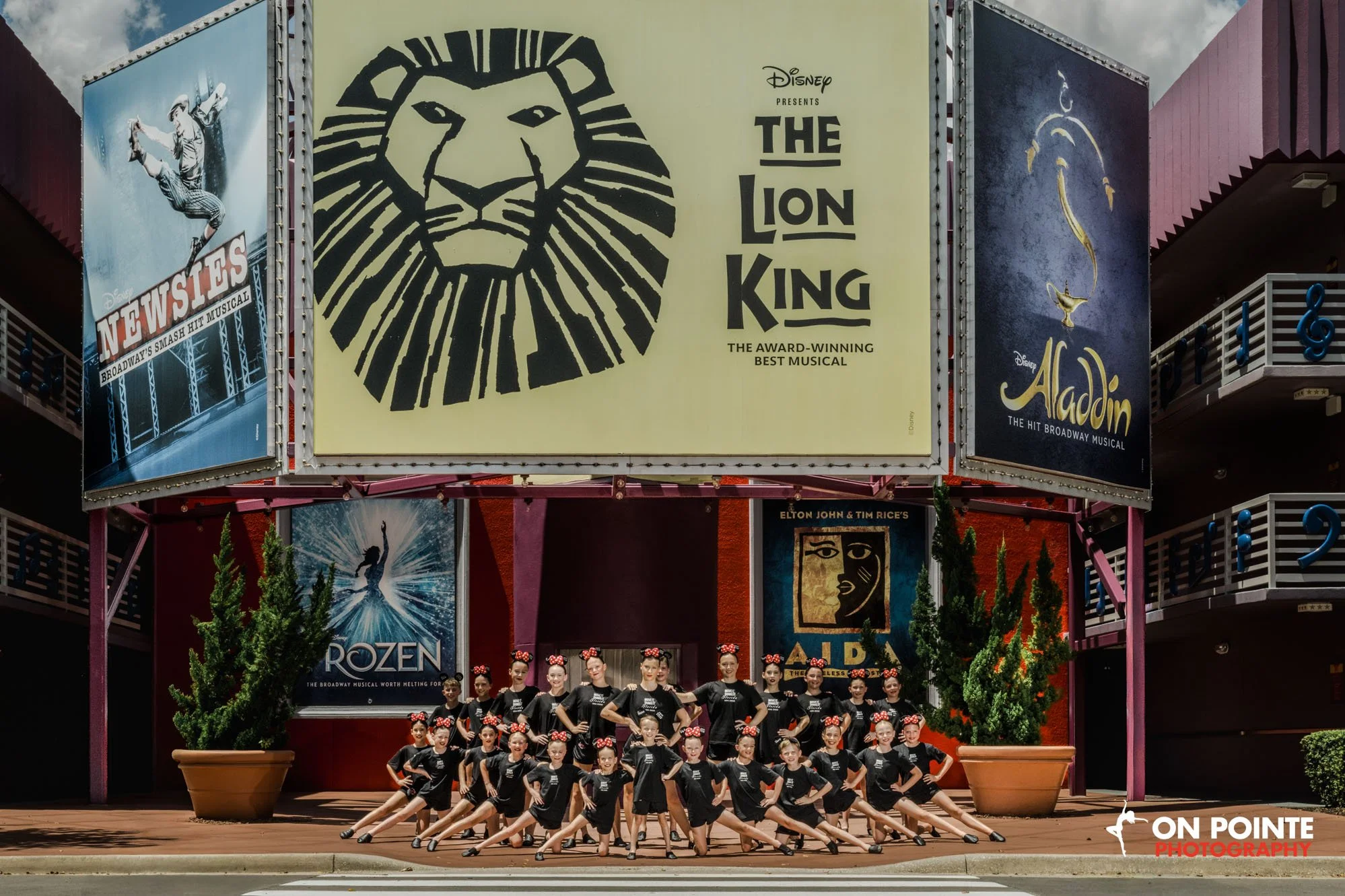 A group of young girls performing a dance routine on an outdoor stage on a Broadway street at Disney California Adventure Park. They are wearing black shirts and black shorts with red and white polka-dot headbands. Large promotional banners for Broad