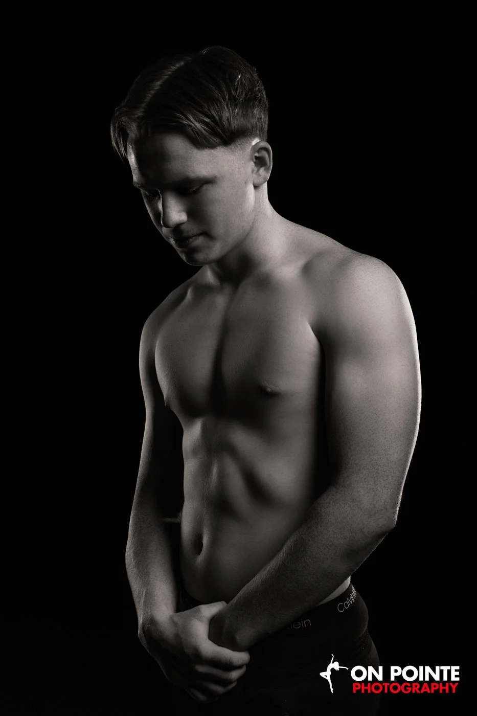 Black and white photo of a shirtless man with head down, hands together against a dark background.