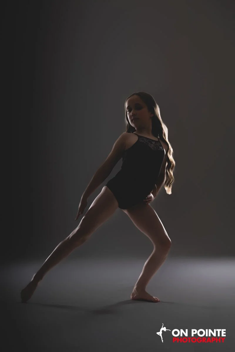 A young girl performing a dance pose, wearing a black leotard, in a studio setting with dark lighting. The logo of 'On Pointe Photography' is visible at the bottom right corner.