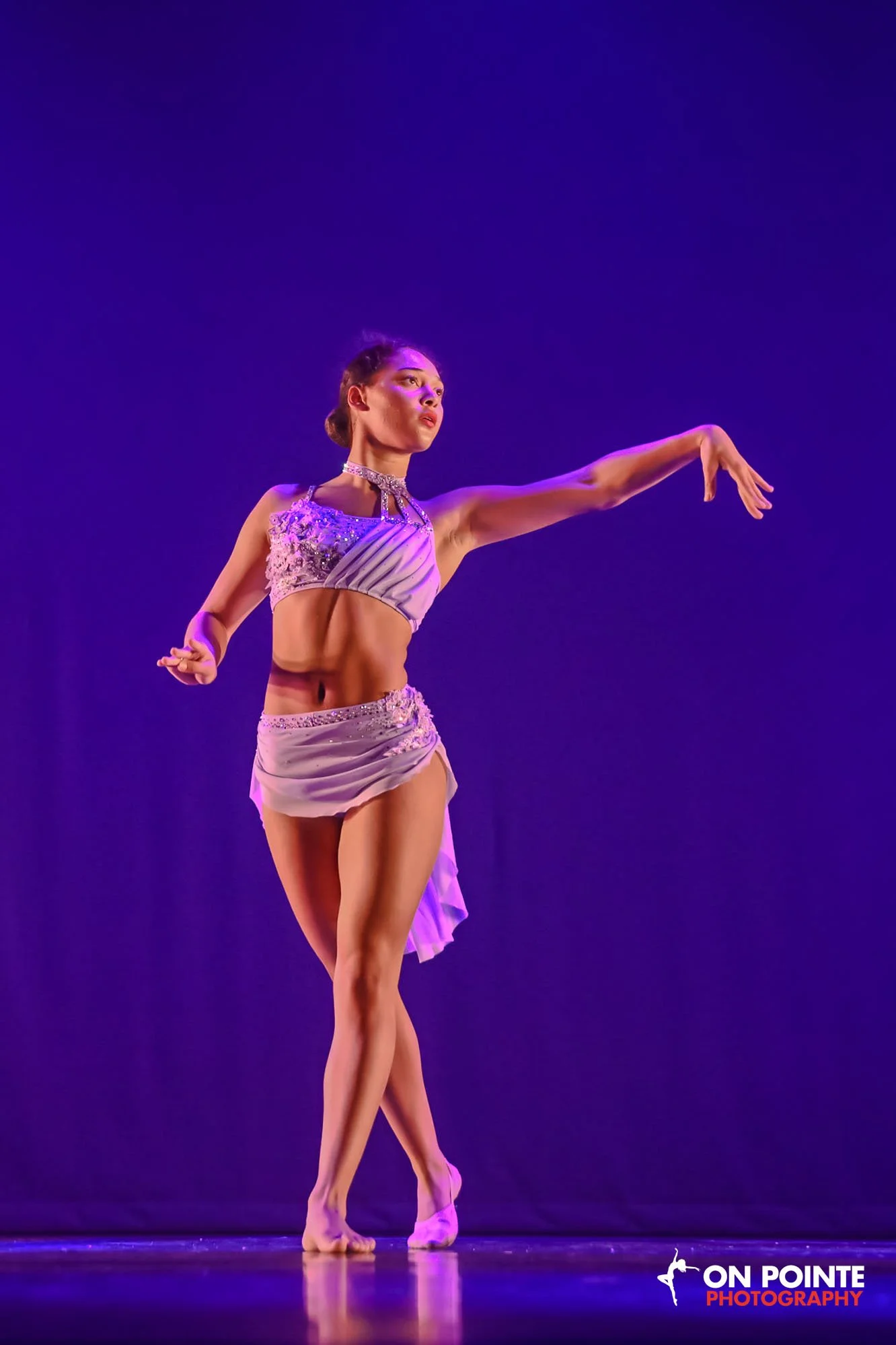 A female ballet dancer performing a pose on stage against a dark purple background. She is wearing a lavender costume with embellishments and a high slit, with her hair styled in a bun. The stage has a reflective surface, and the photo is credited to On Pointe Photography.