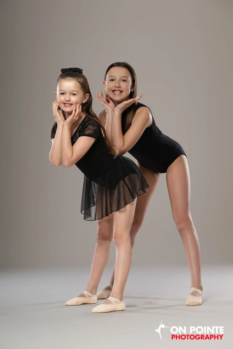 Two young girls in black leotards and ballet slippers posing with their hands under their chins in front of a plain gray background at a dance studio or photo studio.