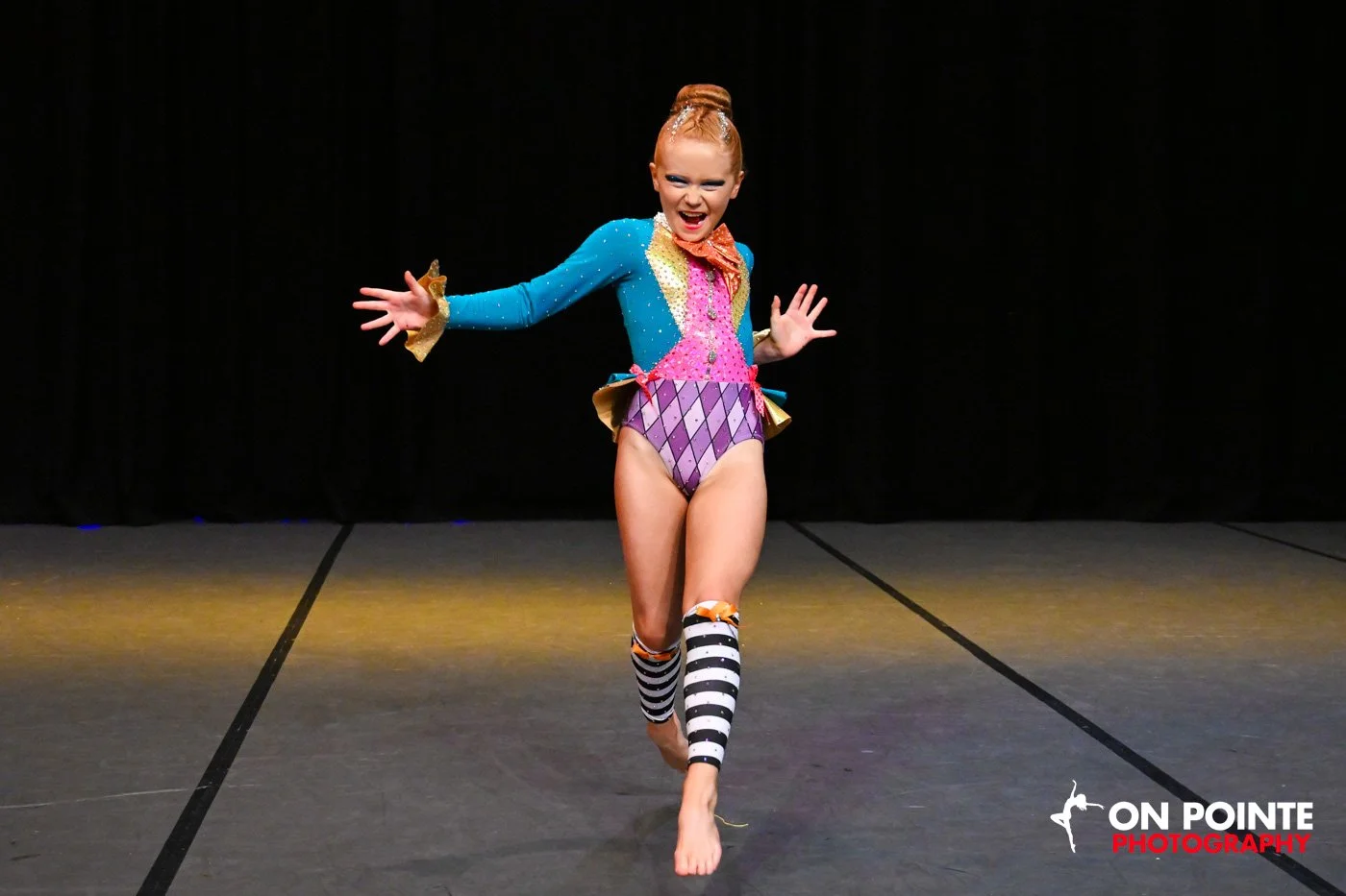 Young girl performing a dance routine in colorful costume with a black background and stage lighting, logo in the bottom right corner.