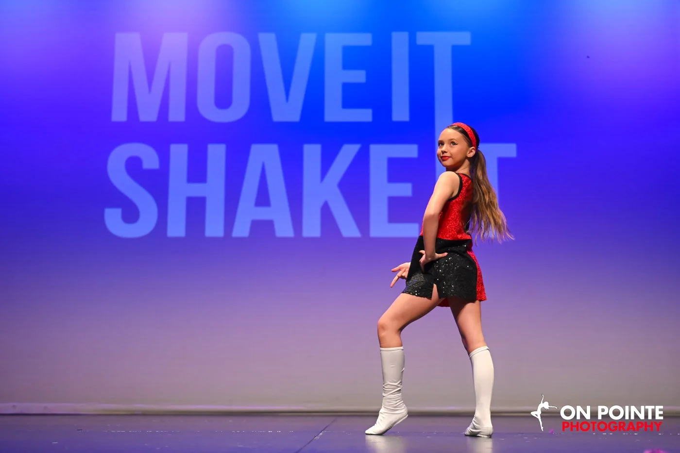 A young girl performing a dance move on stage, with a blue background displaying the words "MOVE IT SHAKE IT" in large letters. She is wearing a red and black costume with white knee-high socks.