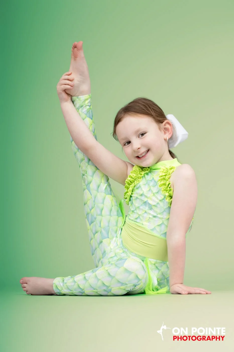 Child in a green dance costume posing with leg raised against a green background.
