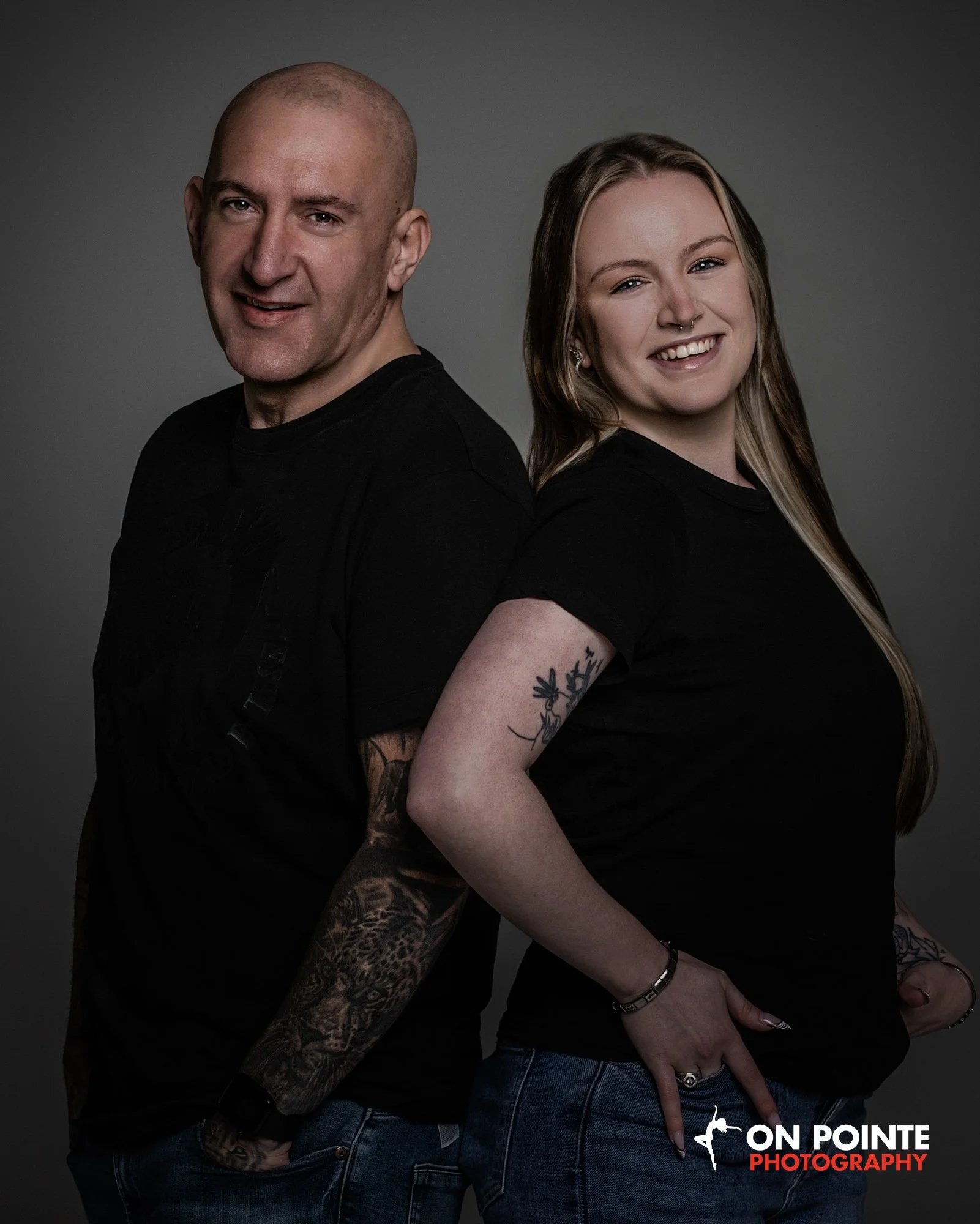 A man and woman stand back to back, smiling, against a dark gray background. The man is bald, wearing a black T-shirt, and has tattoos on his arms. The woman has long blonde hair, a nose piercing, and tattoos on her arms, wearing a black T-shirt and jeans.