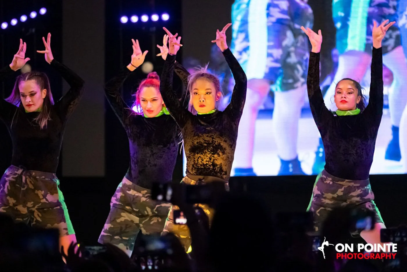 Four female dancers on stage performing a dance routine. They are wearing black velour long-sleeve tops, camouflage shorts, and green neck gaiters. The background features a large screen displaying a reflection of their performance, and there are stage lights shining down.