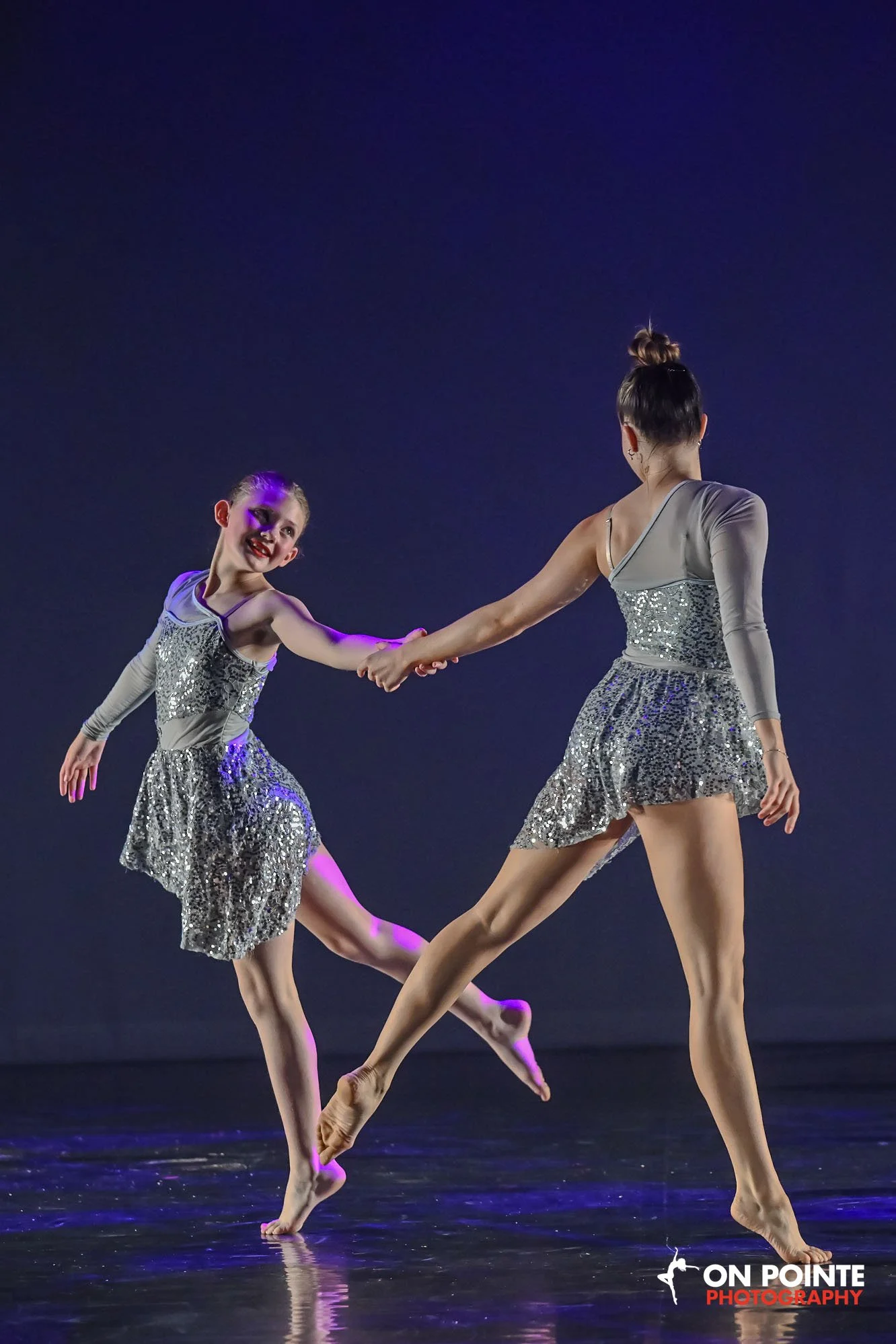 Two young female ballet dancers perform on stage, wearing matching silver sequined dresses, holding hands, and balancing on one leg each, under dark stage lighting with a blue background.