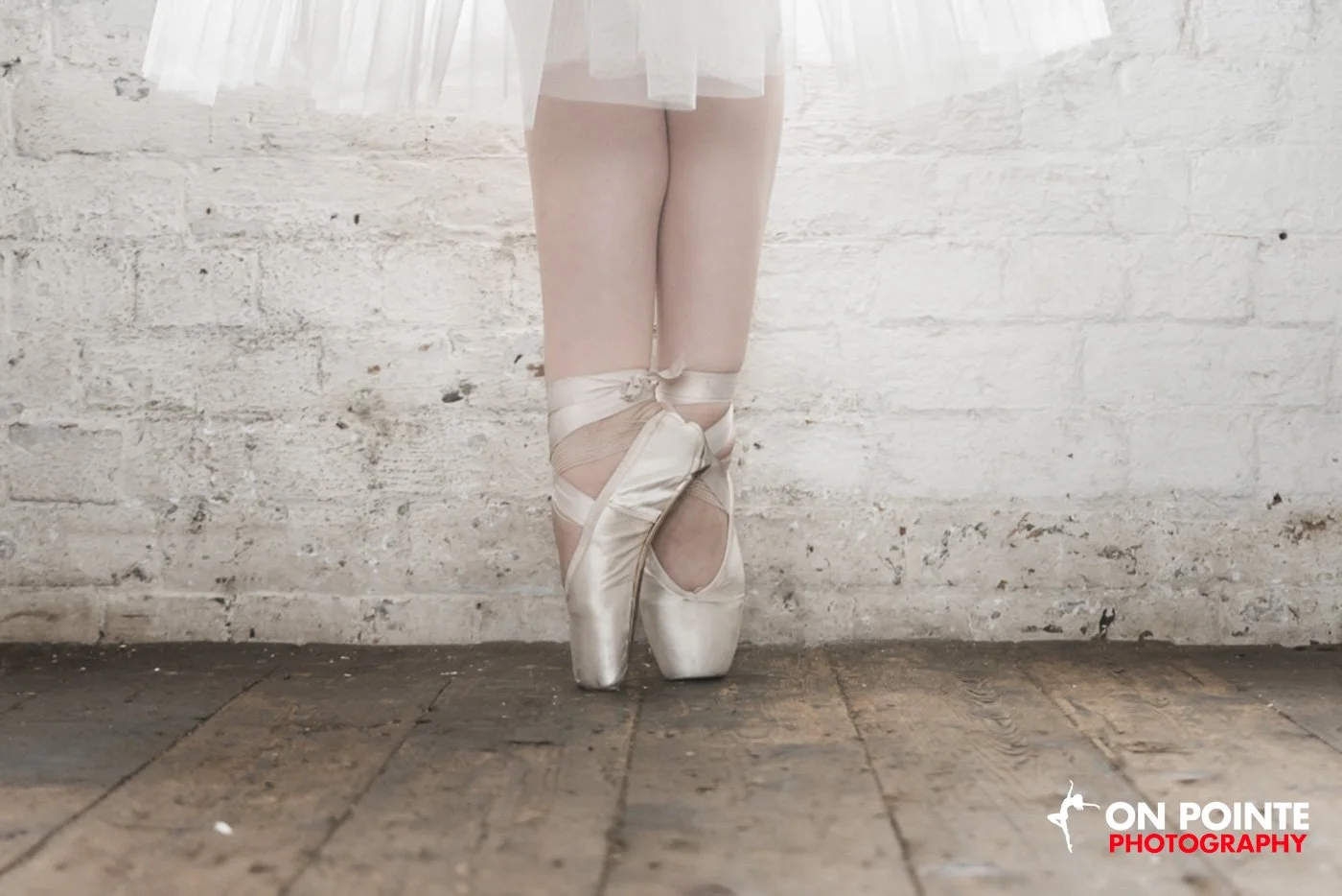 Ballet dancer standing on pointe wearing satin pointe shoes and a white tutu against a brick wall.