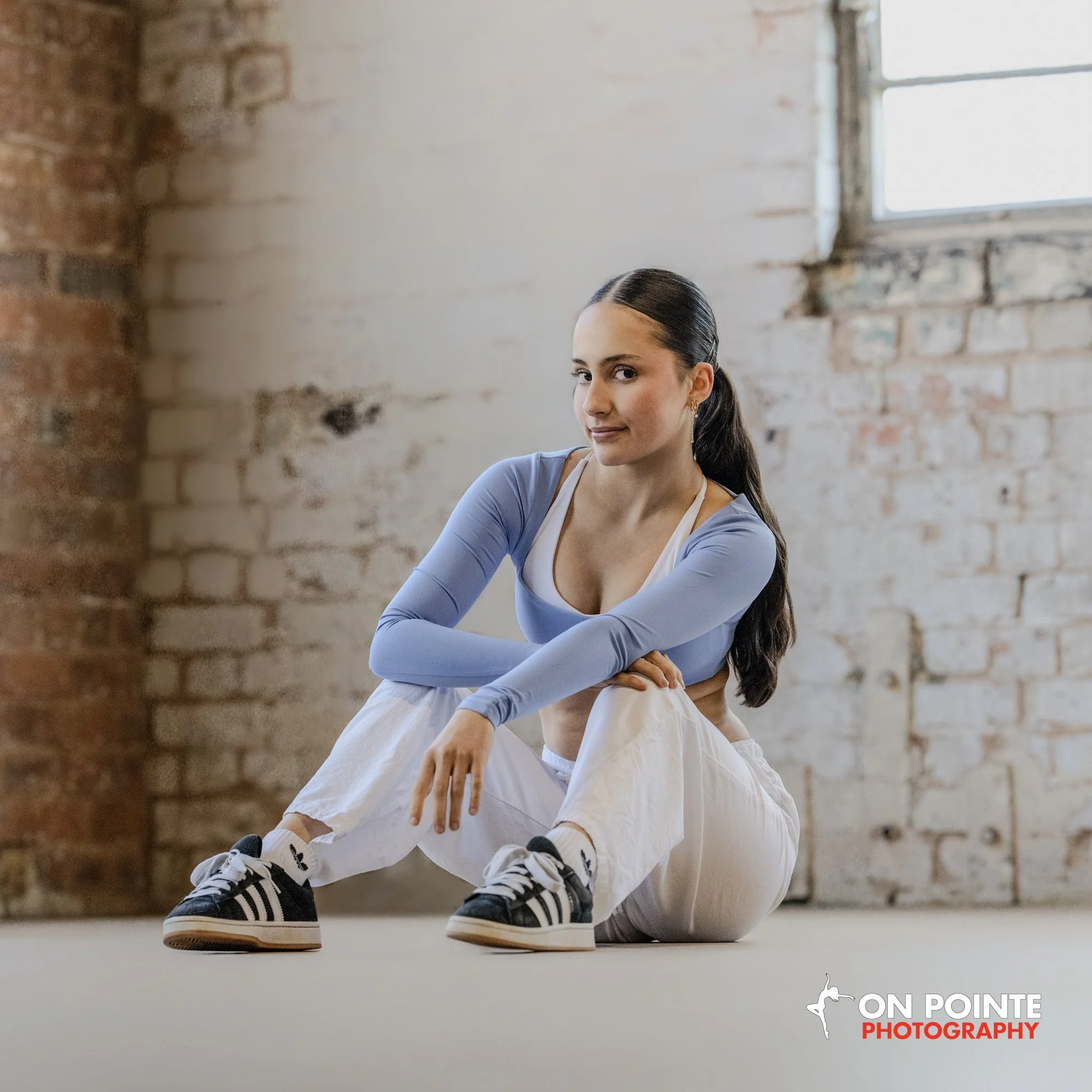 Young woman with long dark hair tied back, wearing a light blue long-sleeve crop top, white pants, and black and white sneakers, sitting on the floor in a room with exposed brick wall and a window, looking at the camera with a slight smile.