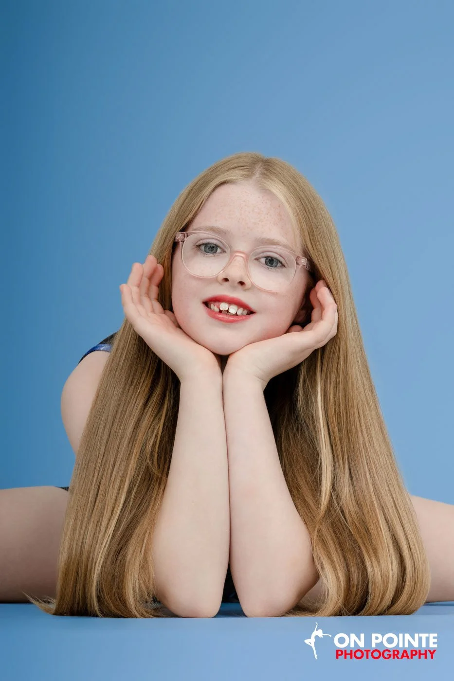 Girl with long blonde hair and glasses posing against a blue background, resting her chin on her hands.