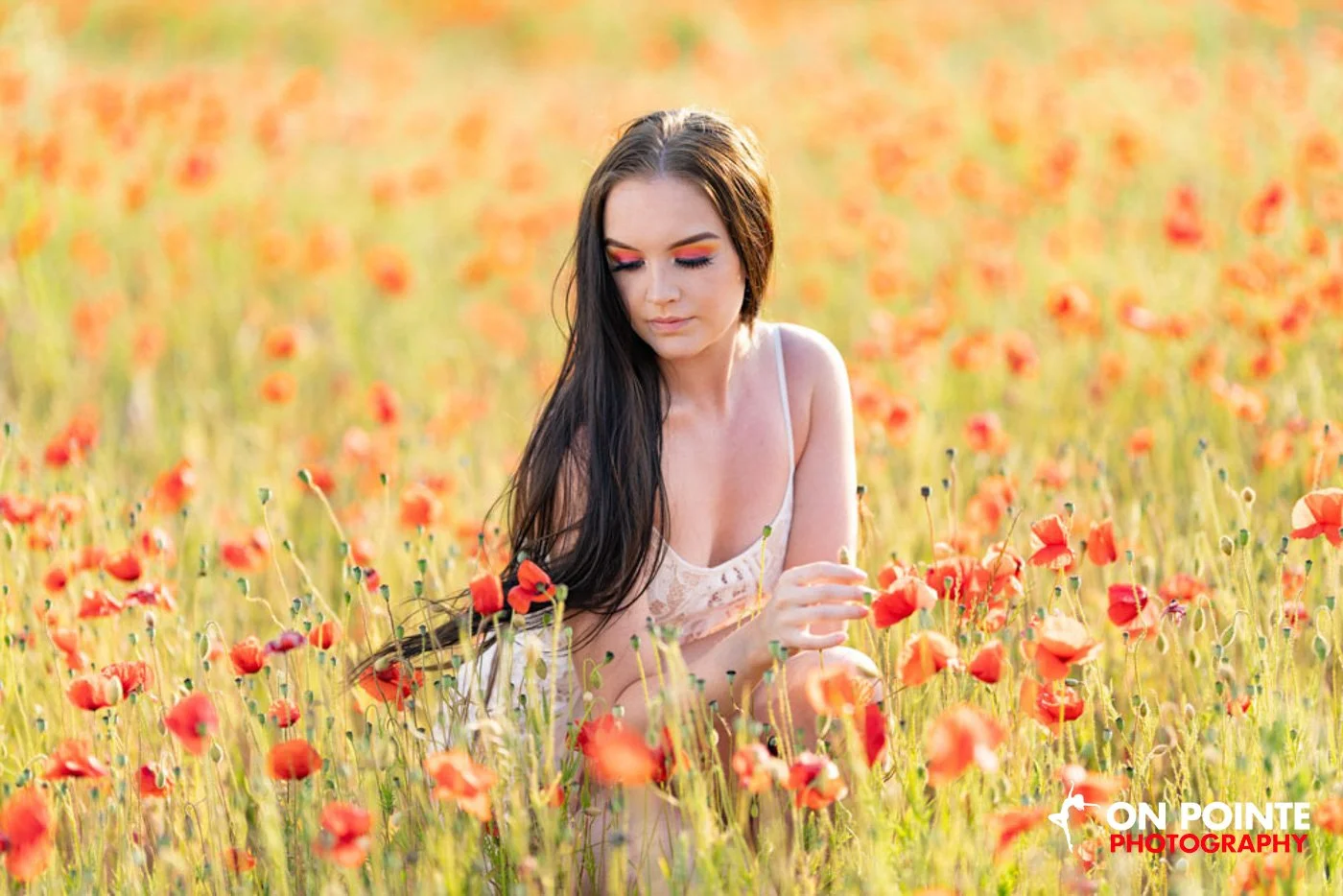 A woman with long hair kneeling in a field of orange poppies under bright sunlight. She is wearing makeup and a light-colored top. The field is densely filled with vibrant poppies.