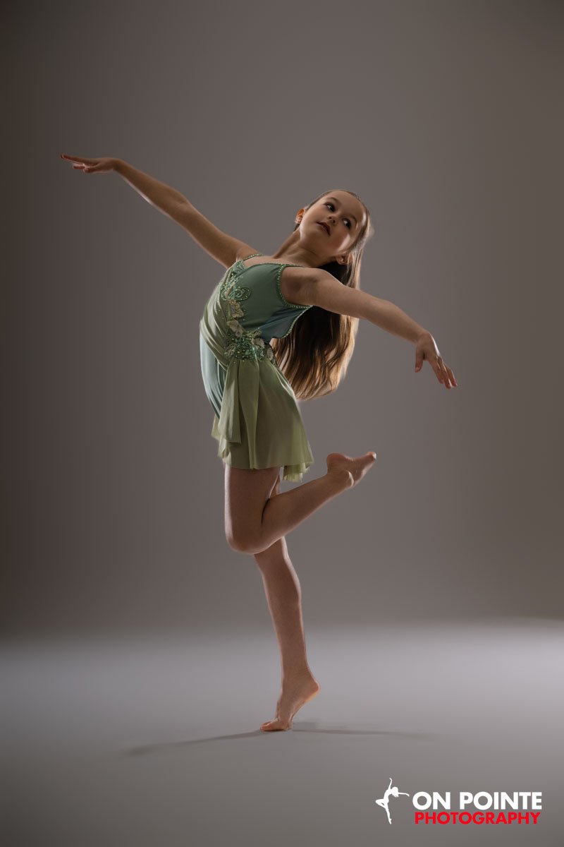 A young girl performing a dance move on a studio floor with a neutral background, wearing a green dress.