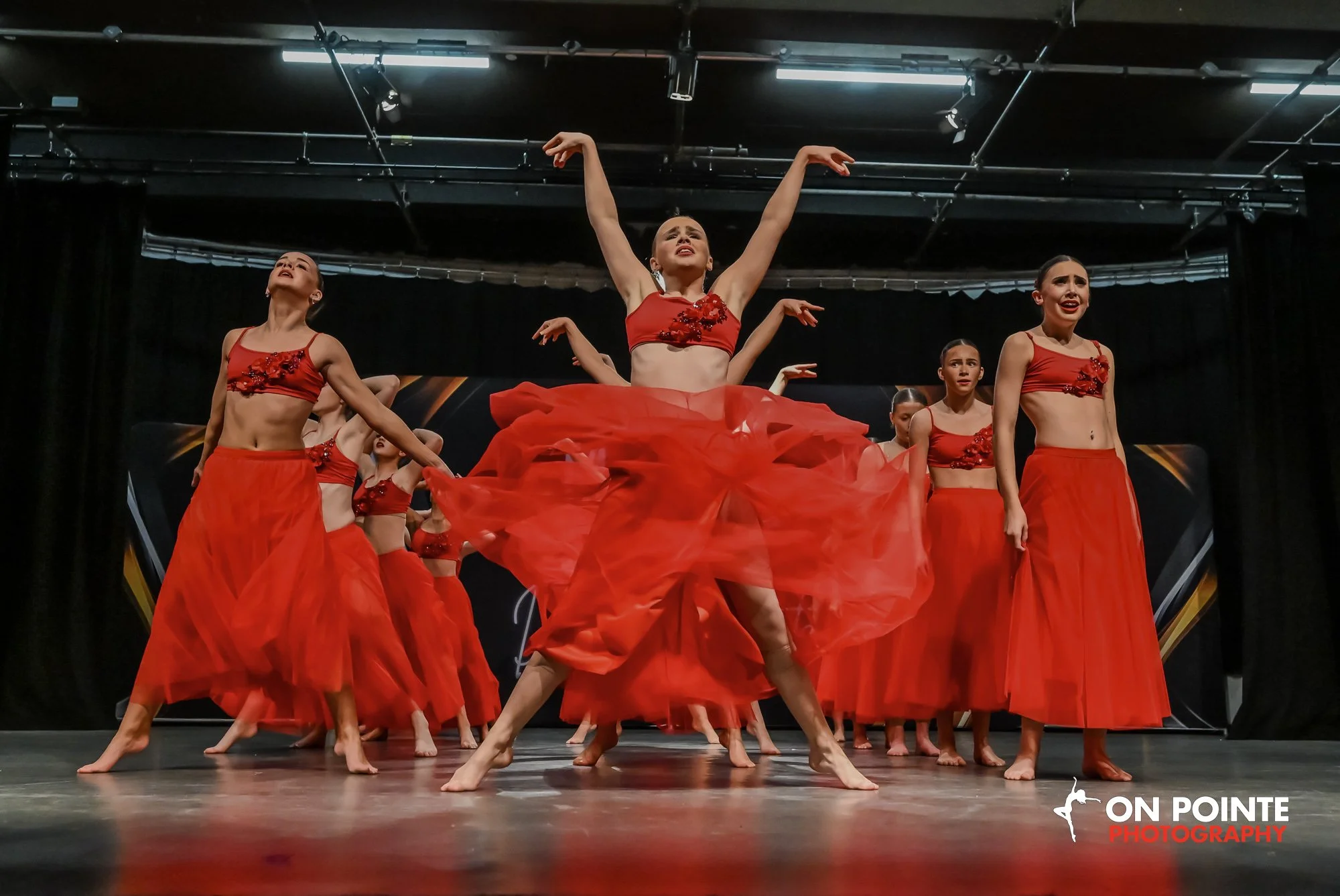 A group of female dancers performing a dance routine on stage, dressed in red costumes with flowing skirts and floral embellishments, with one dancer in the center raising her arms dramatically.