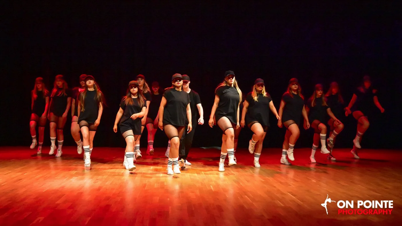 Group of young women dressed in black T-shirts, shorts, and baseball caps performing a dance on a stage with wooden floor, illuminated by red and orange lighting.