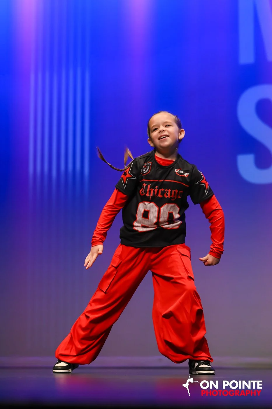 Young girl dancing on stage in a Chicago sports jersey and red pants, smiling with hands on her hips against a purple and blue background.