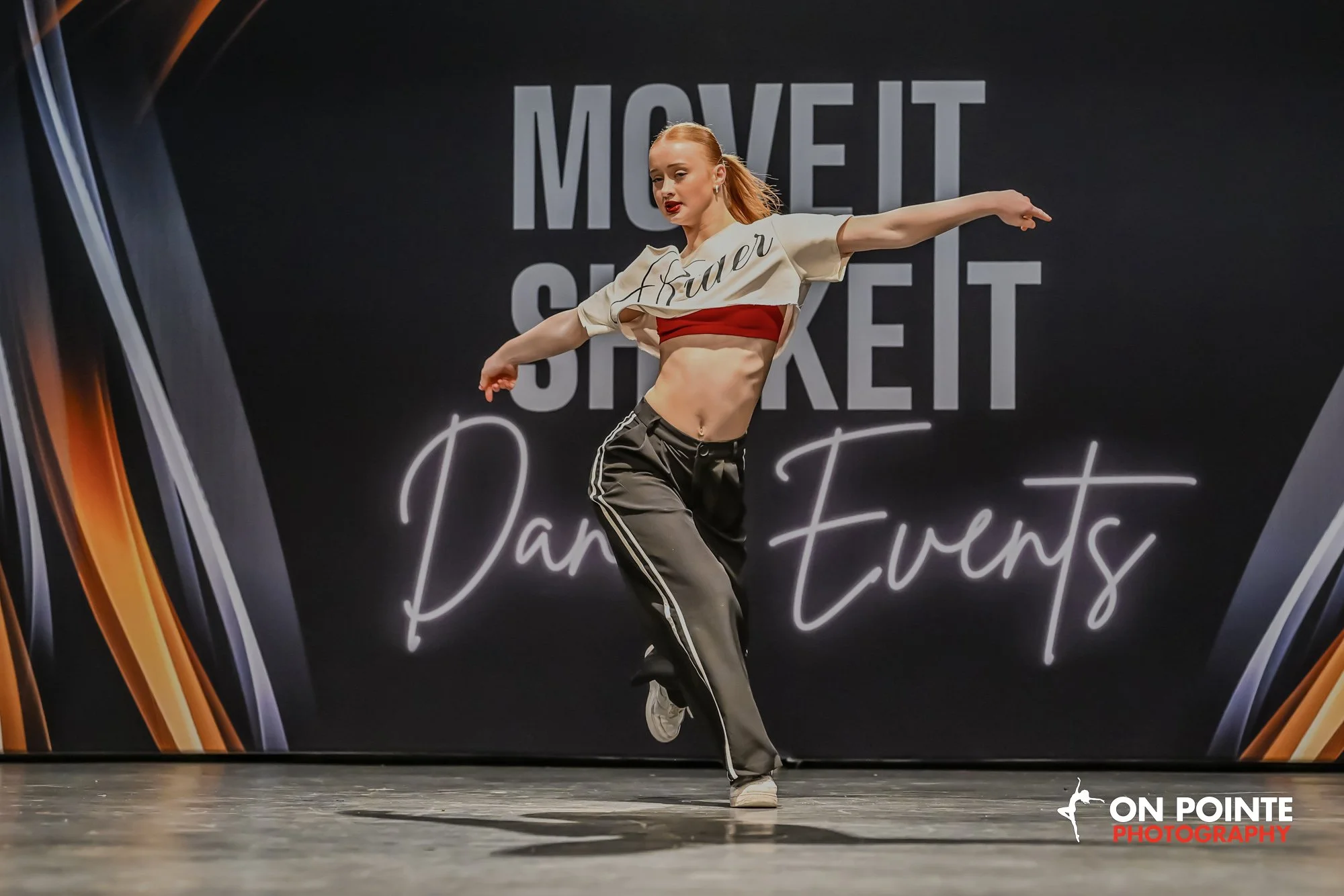 A young female dancer performing on stage at the Move It South Carolina Dance Events competition, wearing a beige and red crop top and black track pants, with a black backdrop featuring the event's name.