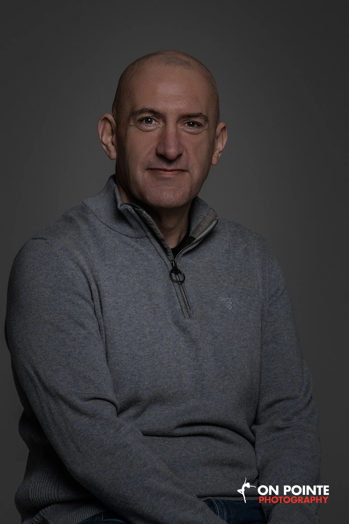 Portrait of a middle-aged man with a bald head, wearing a gray zip-up sweater, sitting against a dark gray background, looking directly at the camera.