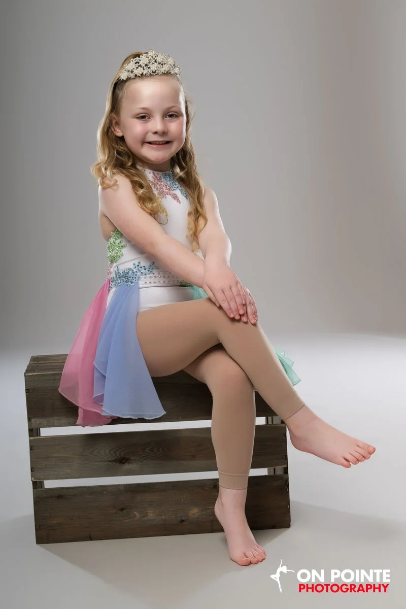 A young girl dressed in a colorful tutu-like dress with a floral headband, sitting on a wooden crate, smiling, with her legs crossed and arms resting on her knee, in a photo studio setting.