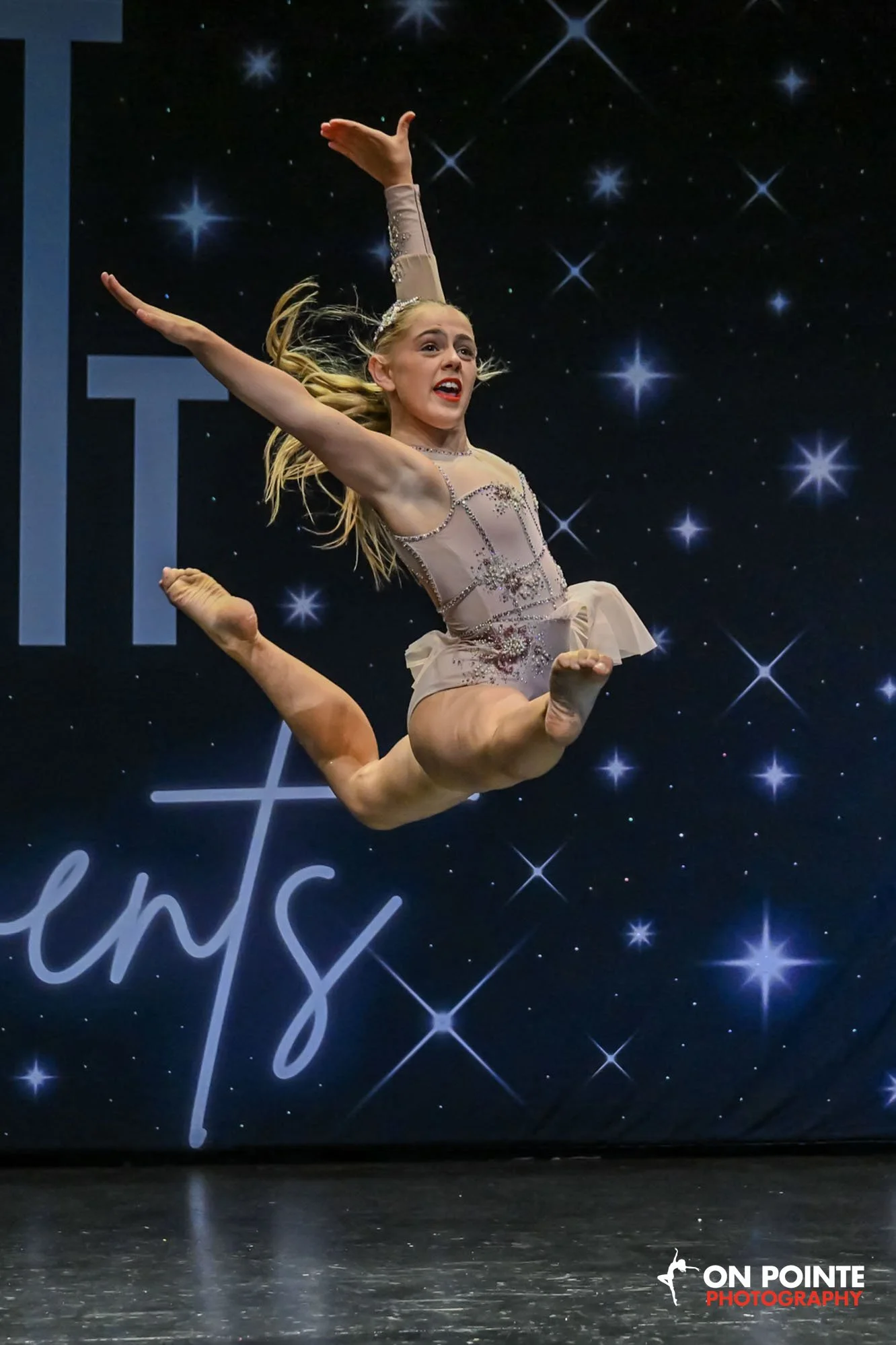 A young female gymnast in a beige and rhinestone leotard performs an acrobatic jump on stage with a starry background and the words "On Pointe Photography" in the bottom right corner.