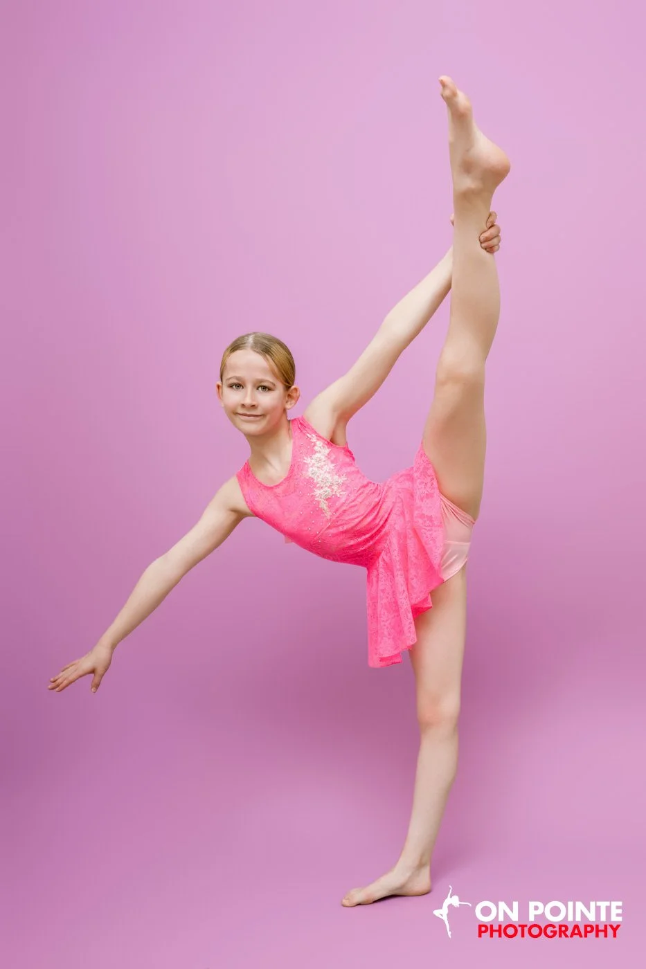 Young dancer in pink costume performing a high leg lift against a pink background.
