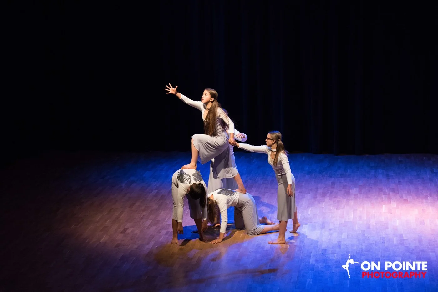 Group of five young women performing a balancing act on stage, with one woman standing on two others' backs, reaching out with her arm, on a dimly lit wooden stage, with a black curtain backdrop, and the logo 'On Pointe Photography' at the bottom rig