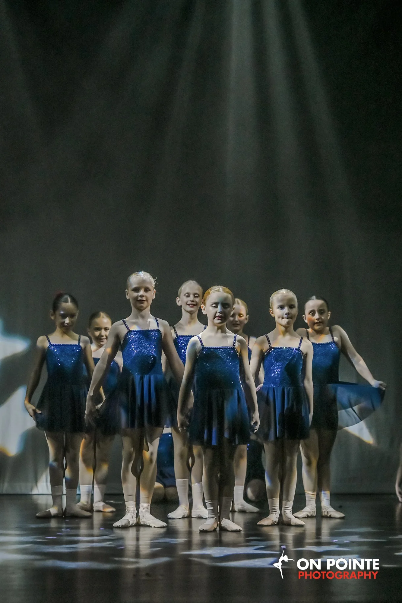 A group of young ballet dancers in blue costumes standing on stage with spotlight shining down.