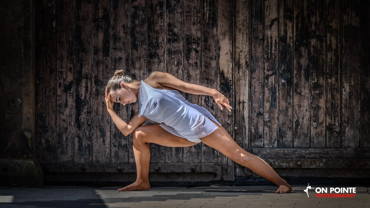 A woman practicing yoga in a low lunge pose against a dark wooden wall, wearing a light gray sleeveless dress.