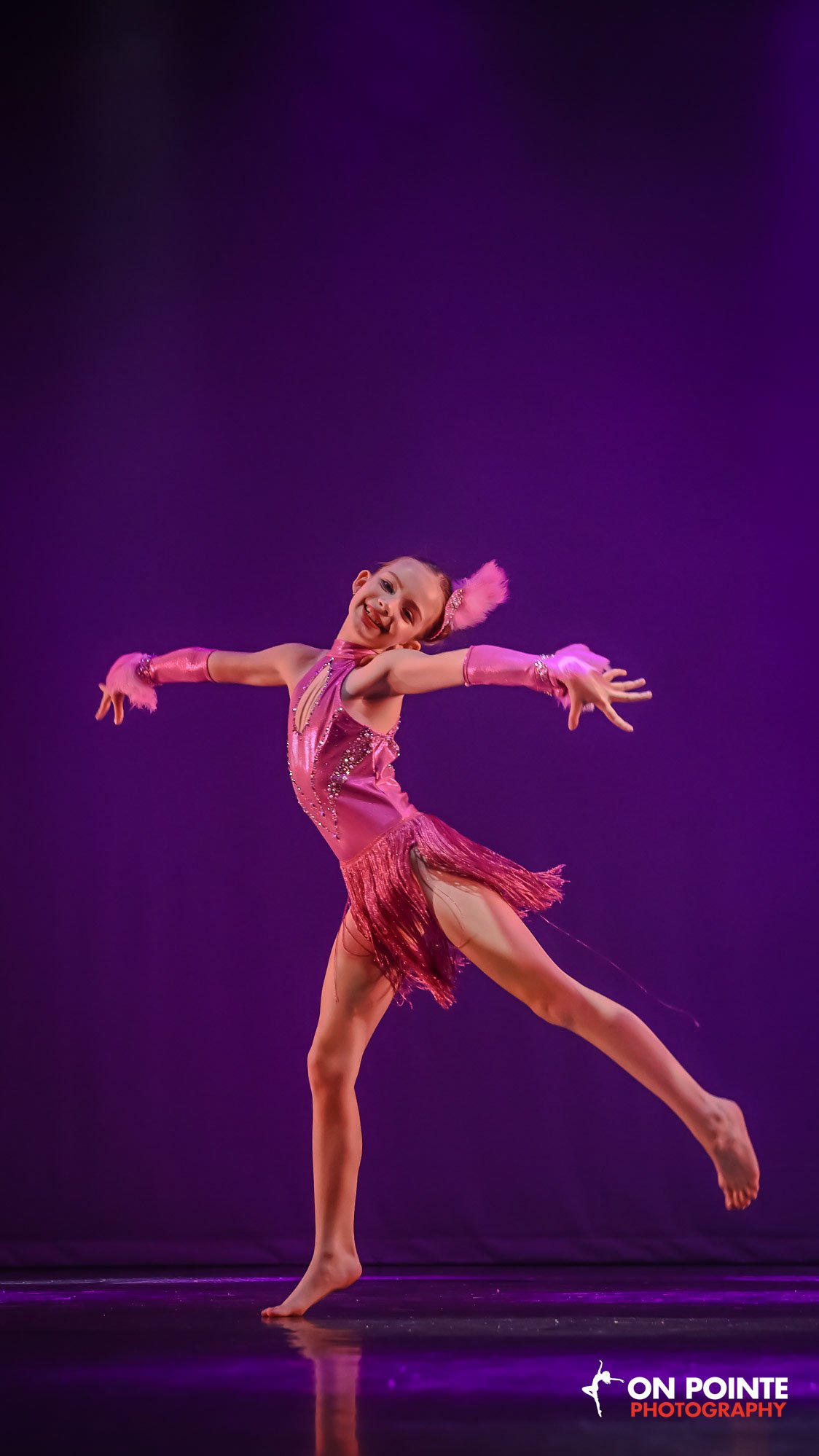 Young girl performing a dance move on stage wearing a pink costume with fringe and matching gloves, with a purple background.