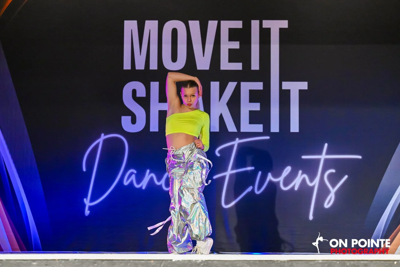 Dancer performing on stage during "Move It Shake It Dance Events" with a neon background.