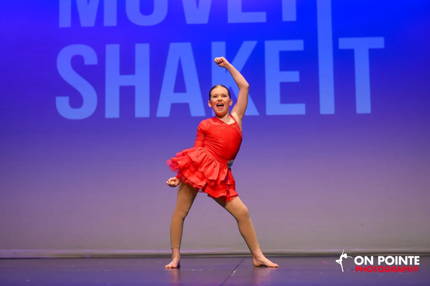 Young girl in a red dance costume striking a pose on stage, with a blue backdrop displaying the words "MOVE, SHAKET."