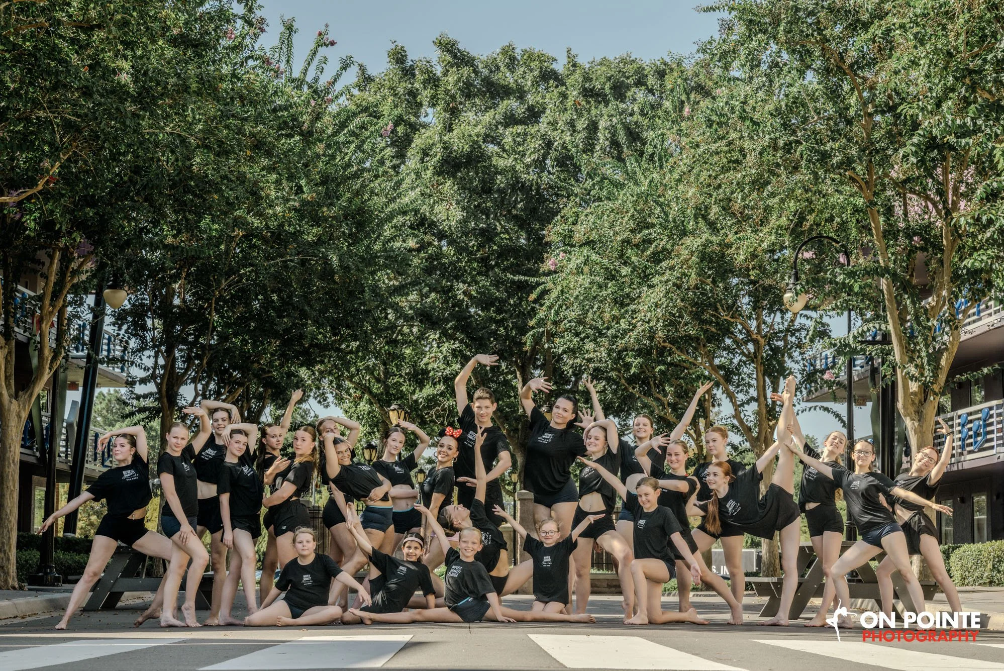 A large group of young dancers in matching black outfits, many with their hair in buns, practicing ballet outside under lush green trees. They are posed in various ballet stances, some standing on one leg, others in pliés or poses, with different arm