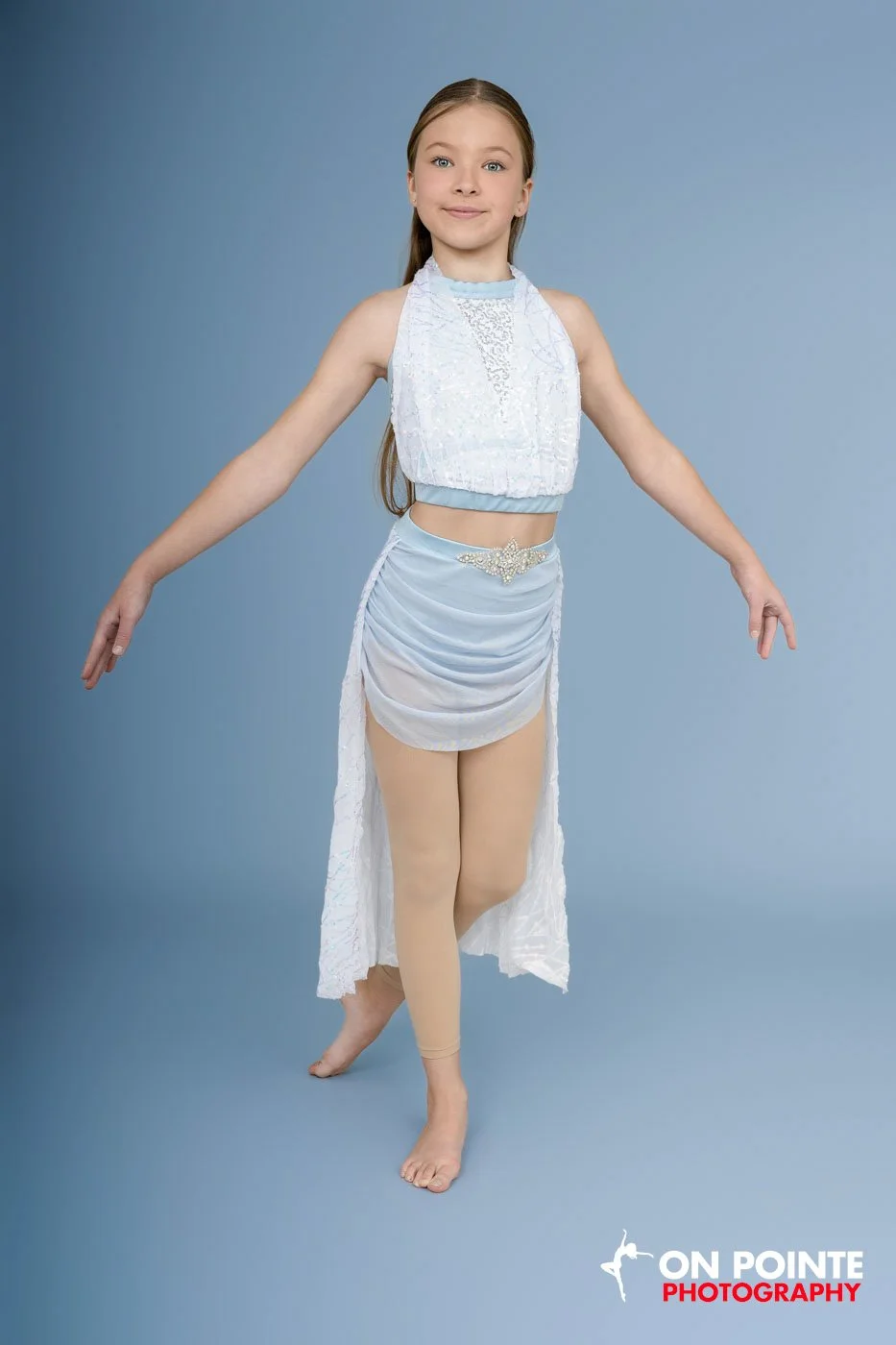 Young girl in a blue dance costume standing in a ballet pose against a blue backdrop.