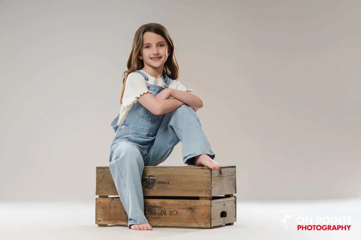 Smiling young girl with long brown hair sitting on wooden crates against a plain beige background, wearing a white shirt and denim overalls.