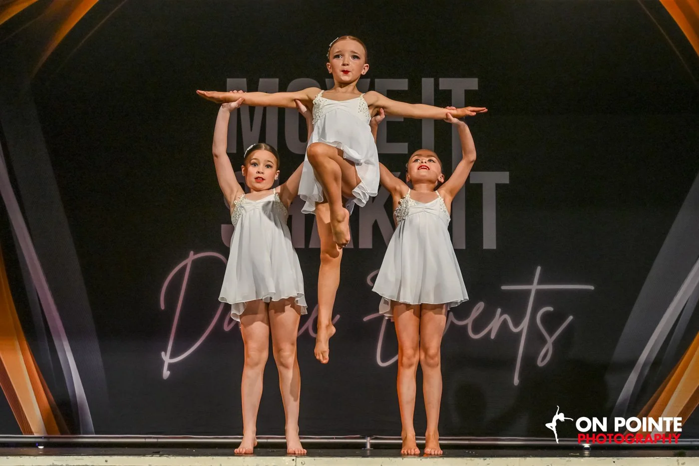 Three young dancers performing on stage, wearing white dresses, with one dancer being lifted by the others.