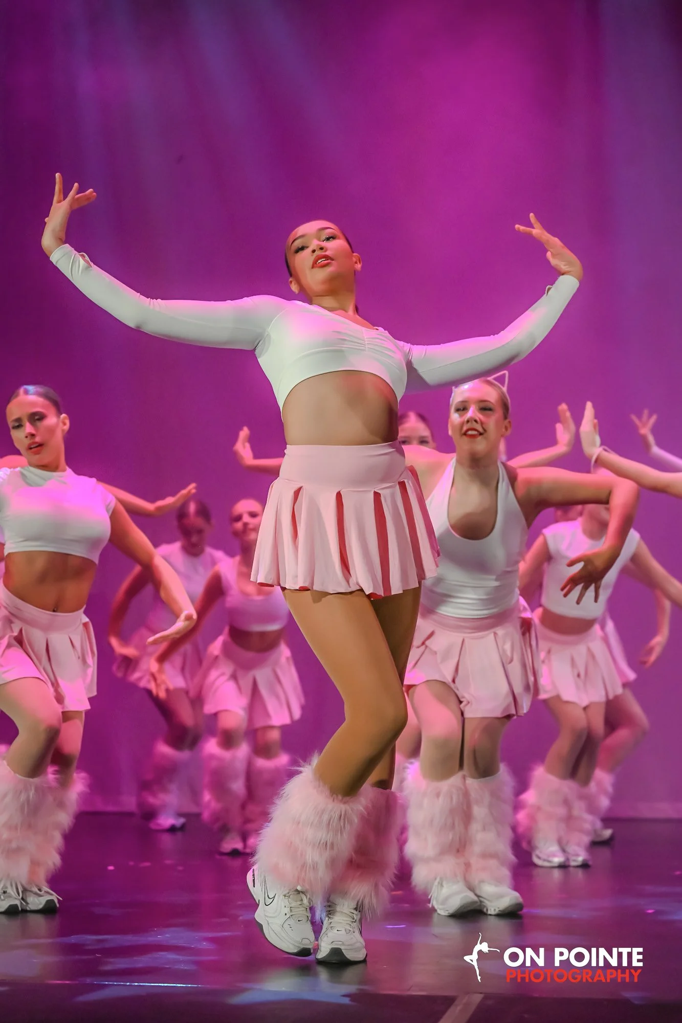 A group of female dancers in pink and white costumes performing on stage with pink lighting.