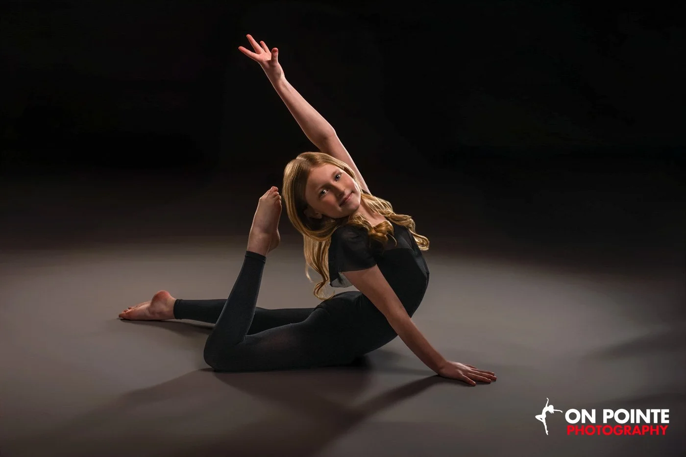 Young girl in a black leotard doing a pose on the floor with one leg bent and arm extended upward, smiling, with "On Pointe Photography" logo in corner.
