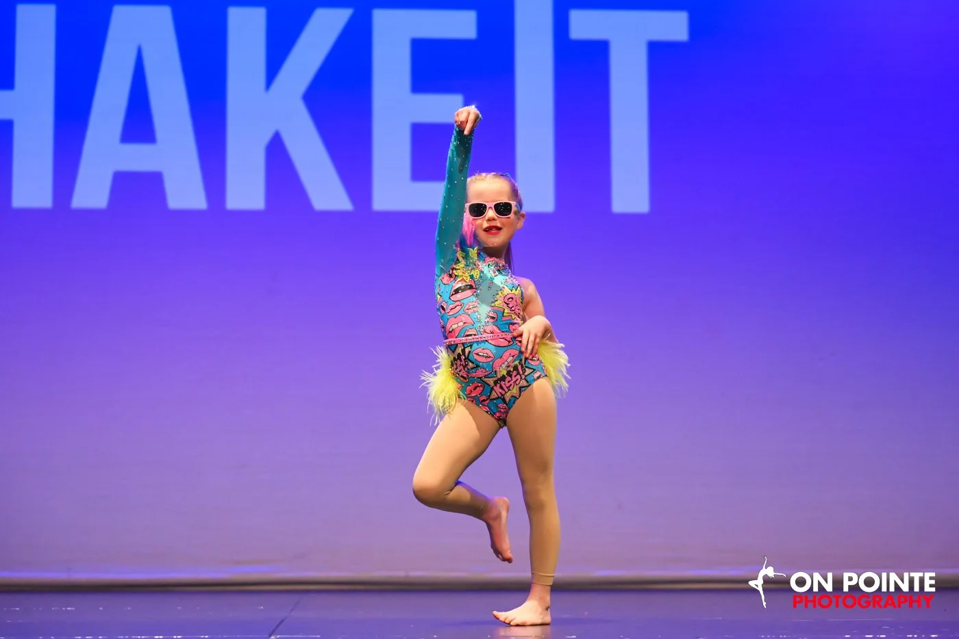A young girl dressed in a colorful, glittery leotard with neon yellow accents, striking a dance pose on stage. She is wearing sunglasses, has her hair tied back, and is balancing on one foot with her other leg bent and raised behind her.