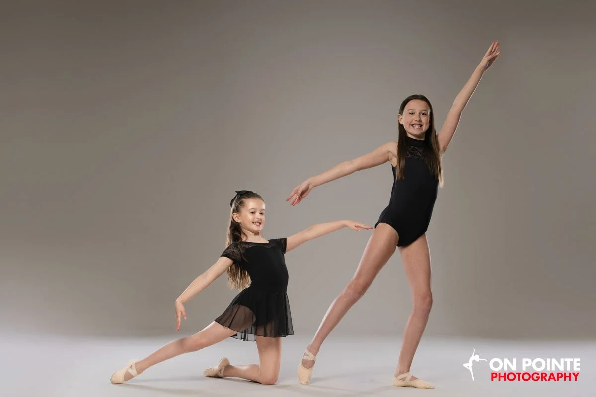 Two young girls in black dance costumes pose gracefully in a dance studio with a neutral backdrop.