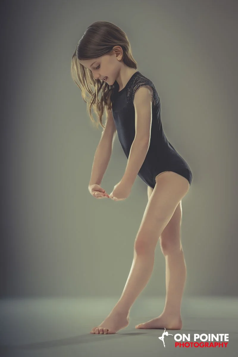 A young girl with long hair in a black lace top and shorts standing on a neutral background, looking down and adjusting her sock.
