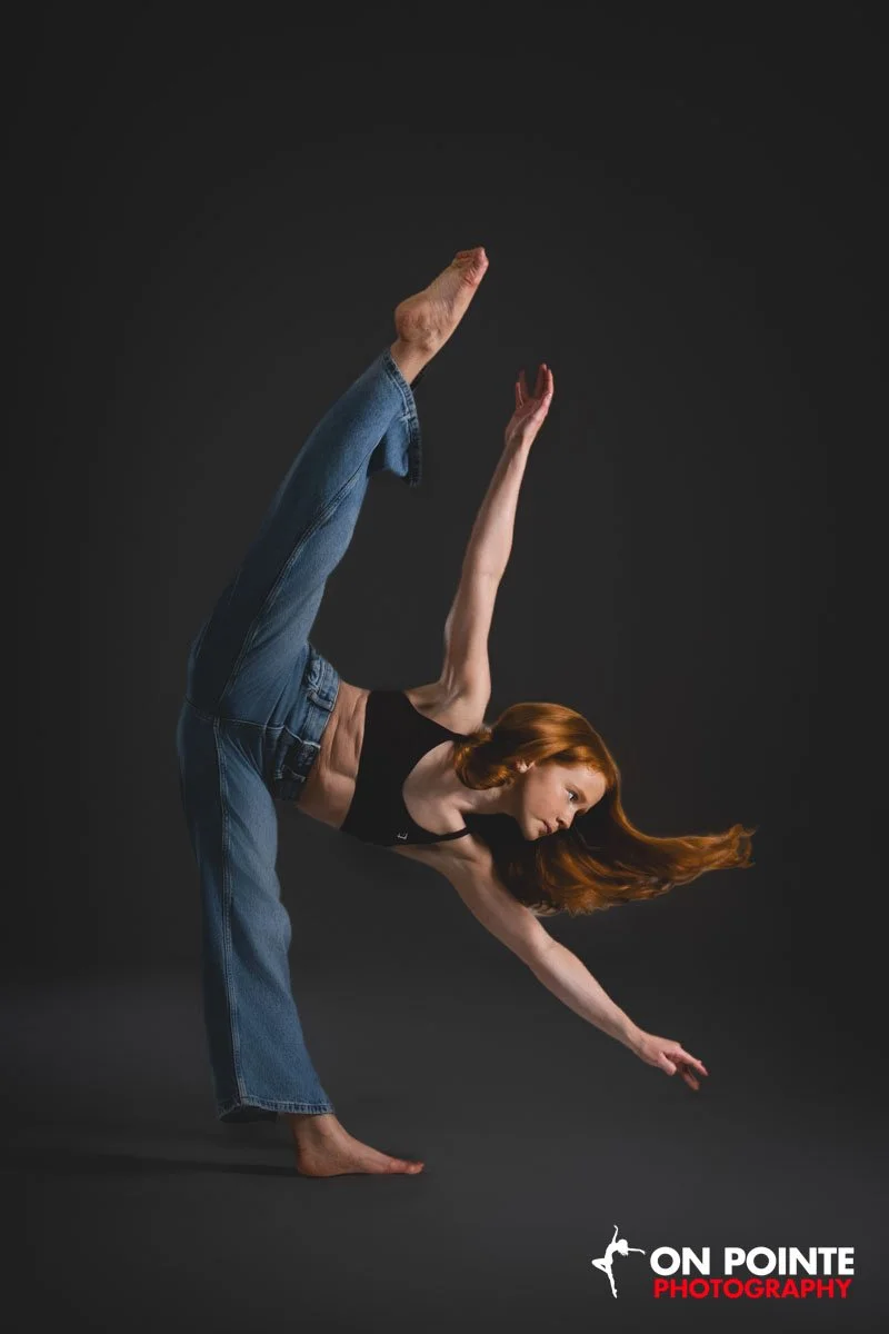 A woman in jeans and a black crop top performing a dance or gymnastic pose against a dark background, with one leg bent and raised, and her body bent forward with one arm extended downward and to the side.
