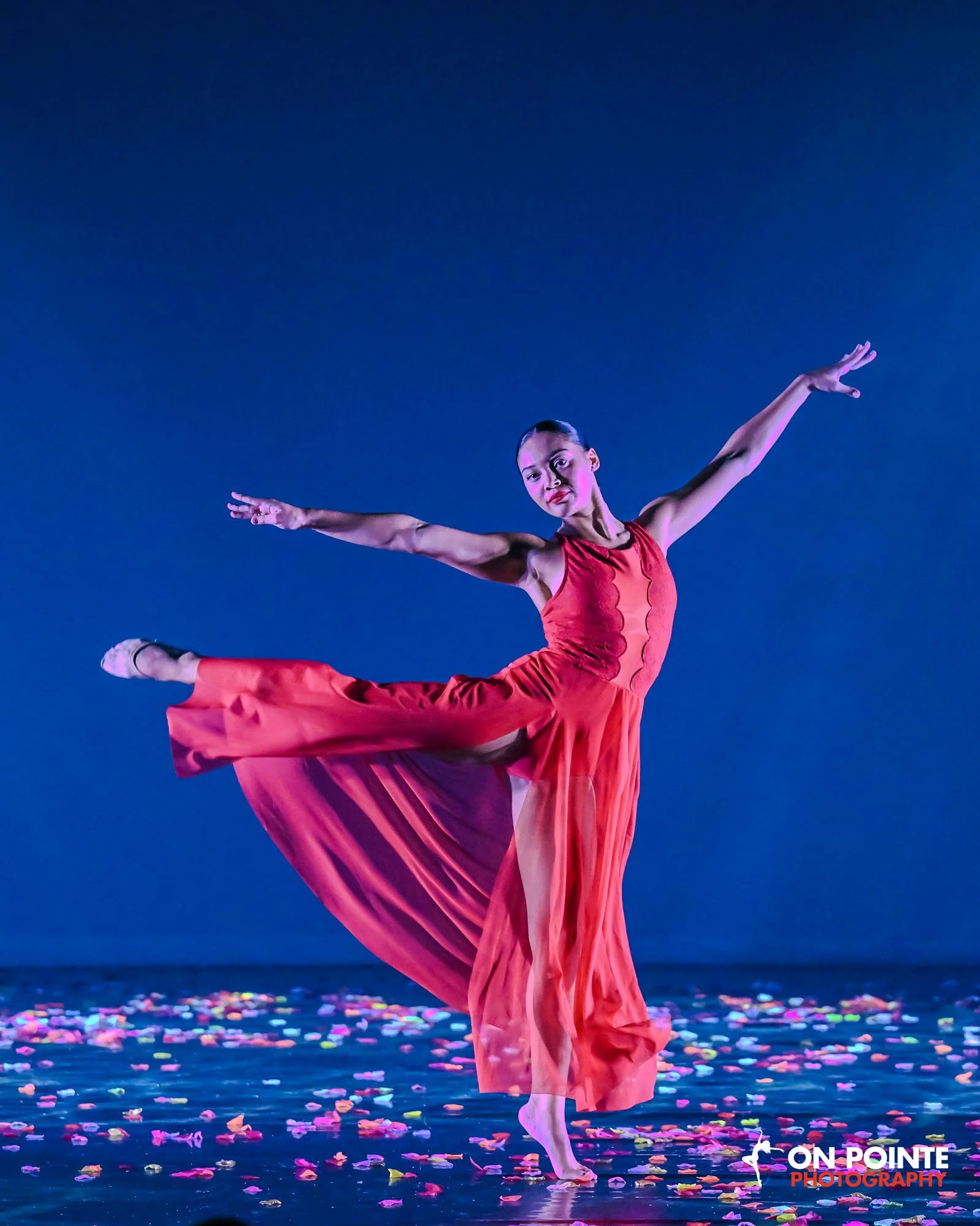 A woman in a red dress performs a ballet dance on stage with pink petals scattered on the floor and a blue background.