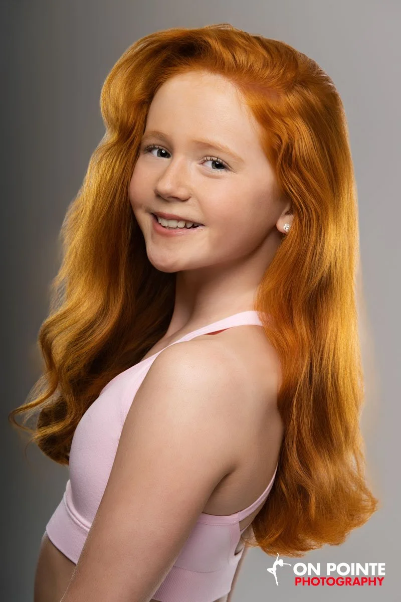 Portrait of a young girl with long red hair, smiling, wearing a pink top, with a gray background.