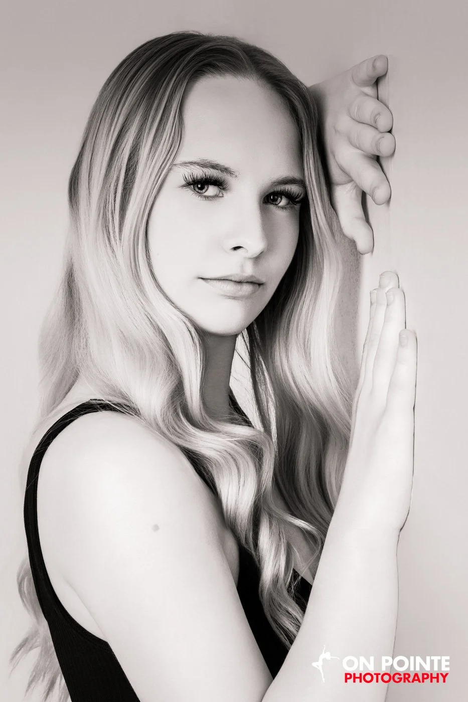 Black-and-white portrait of a woman with long wavy hair, looking at the camera, with her hands posed vertically beside her face.