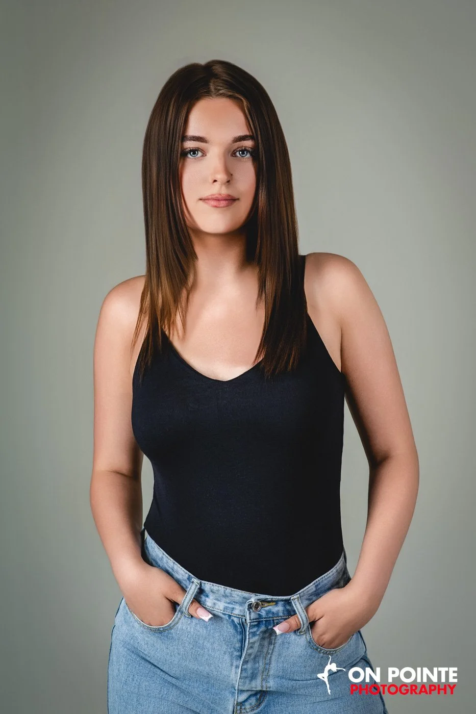 A young woman with straight brown hair, wearing a black tank top and light blue jeans, posing against a plain gray background.